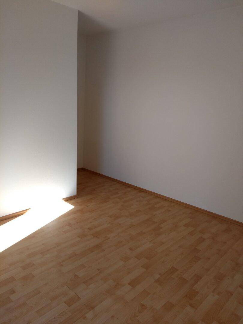 Leerer Raum mit hellem Laminatboden und weiß gestrichenen Wänden, gut beleuchtet.