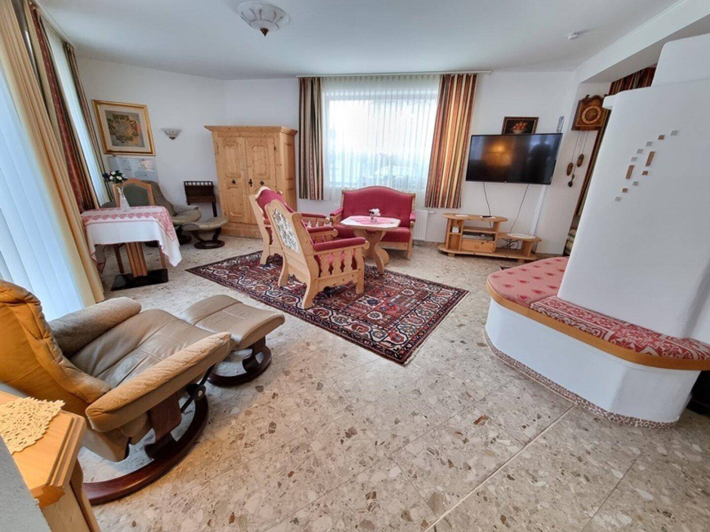 Helles Wohnzimmer mit Kachelofen, bequemen Sitzgelegenheiten und Blick nach draußen.