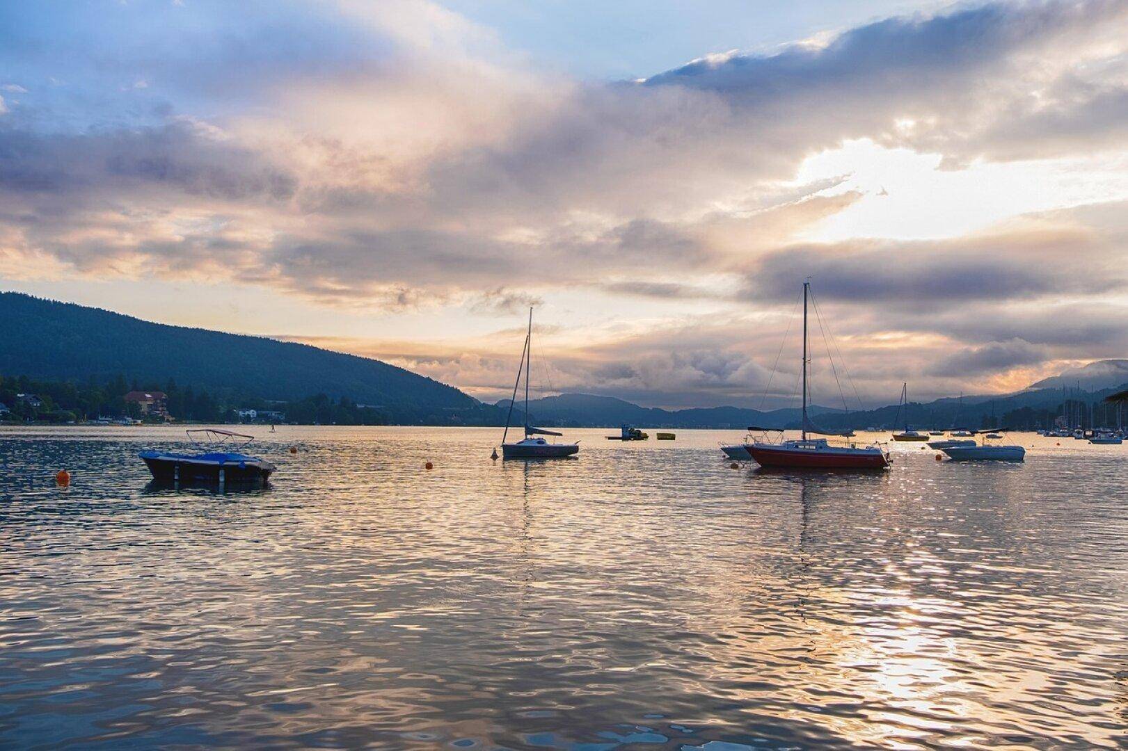 Ruhiger Sonnenuntergang über einem See mit Segelbooten und bewaldeten Bergen im Hintergrund.