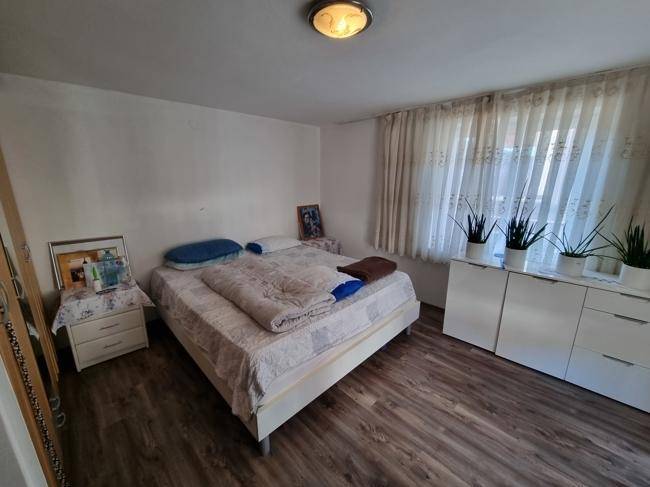 Schlafzimmer mit Doppelbett, Holzoptikboden und hellen Vorhängen am Fenster.