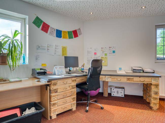 Helles Büro mit großem Schreibtisch aus Holzpaletten, Fenster und Teppichboden.