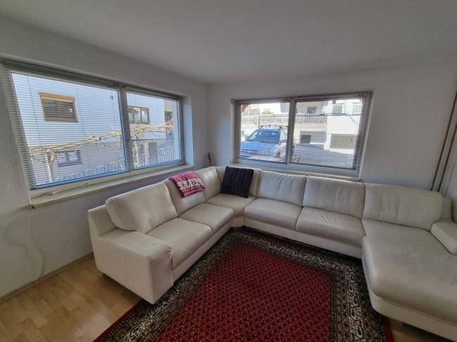 Gemütliches Wohnzimmer mit großem hellen Ecksofa, Teppich und breiten Fenstern.