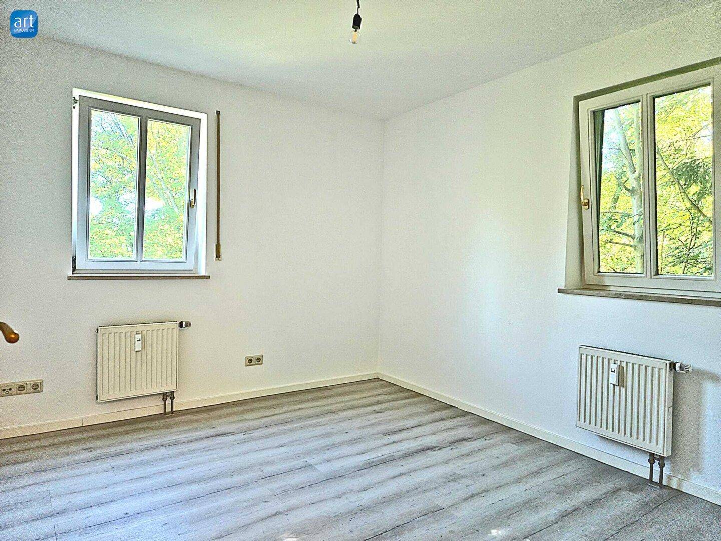 Helles Schlafzimmer mit Laminatboden und zwei Fenstern mit Blick ins Grüne.