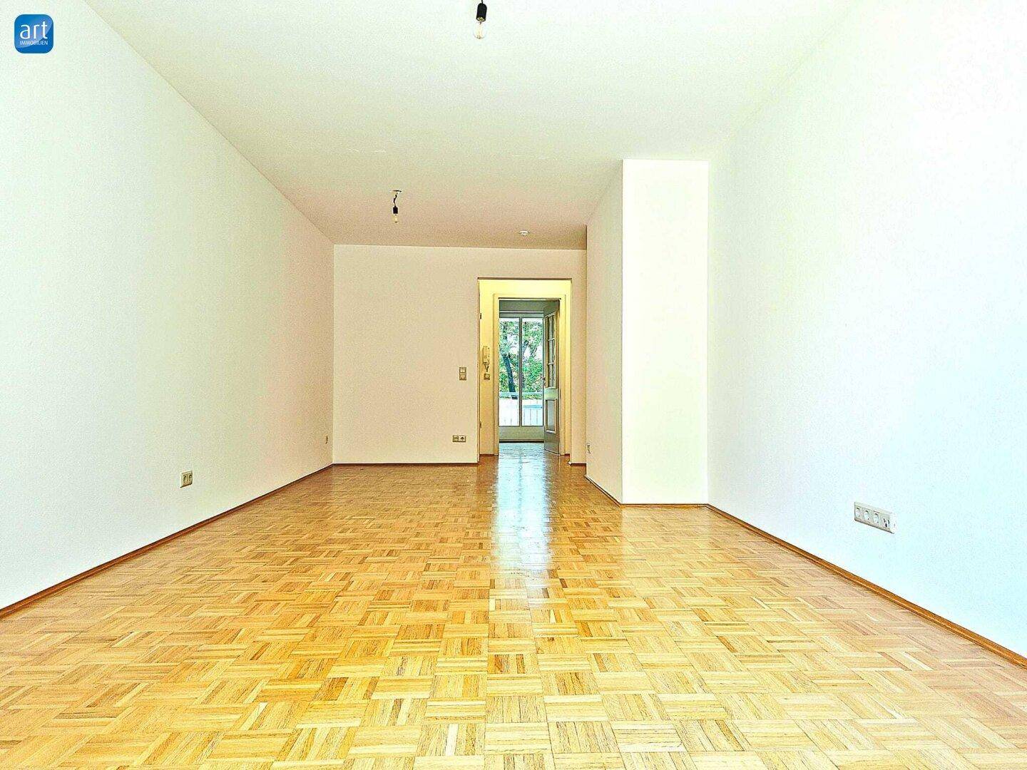 Heller, leerer Wohnbereich mit Parkettboden und Durchgang zu einem weiteren Raum.
