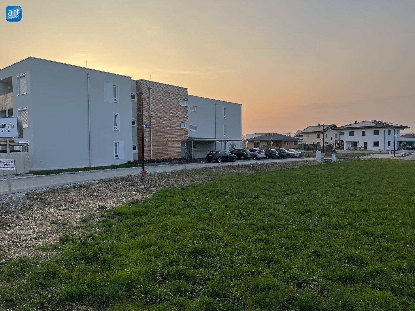Modernes Mehrfamilienhaus mit Holzverkleidung und Parkplätzen in ländlicher Umgebung bei Sonnenuntergang.