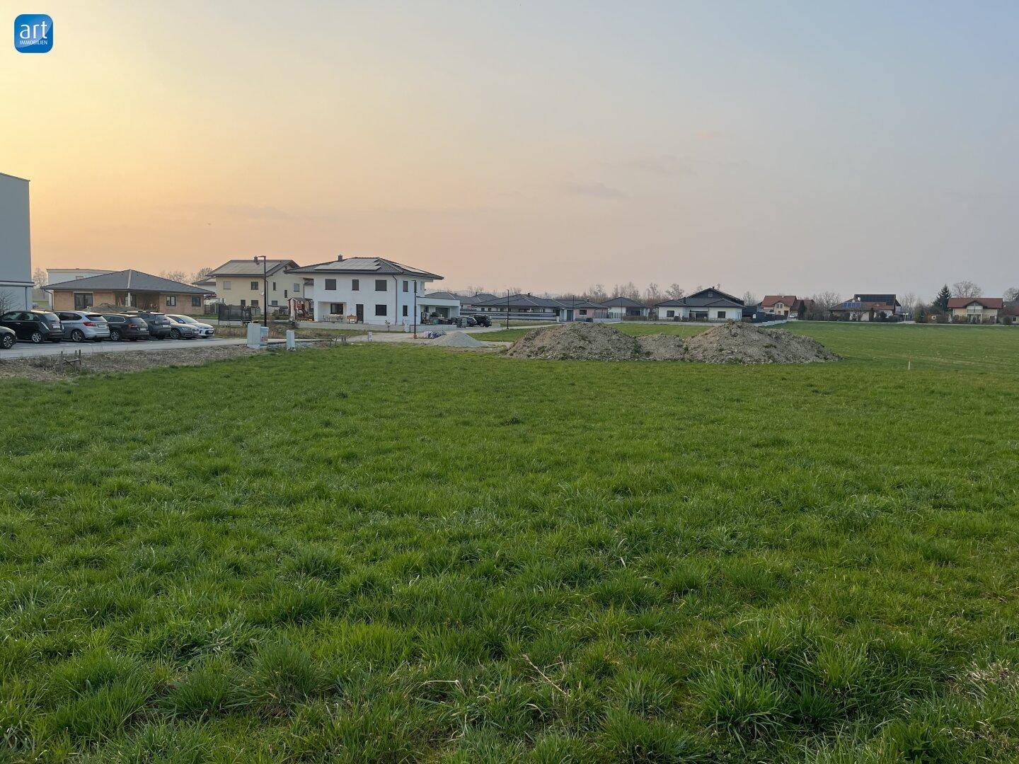 Grüne Wiese mit Erdhügeln und Blick auf angrenzende Wohnhäuser und Parkplätze in der Ferne.