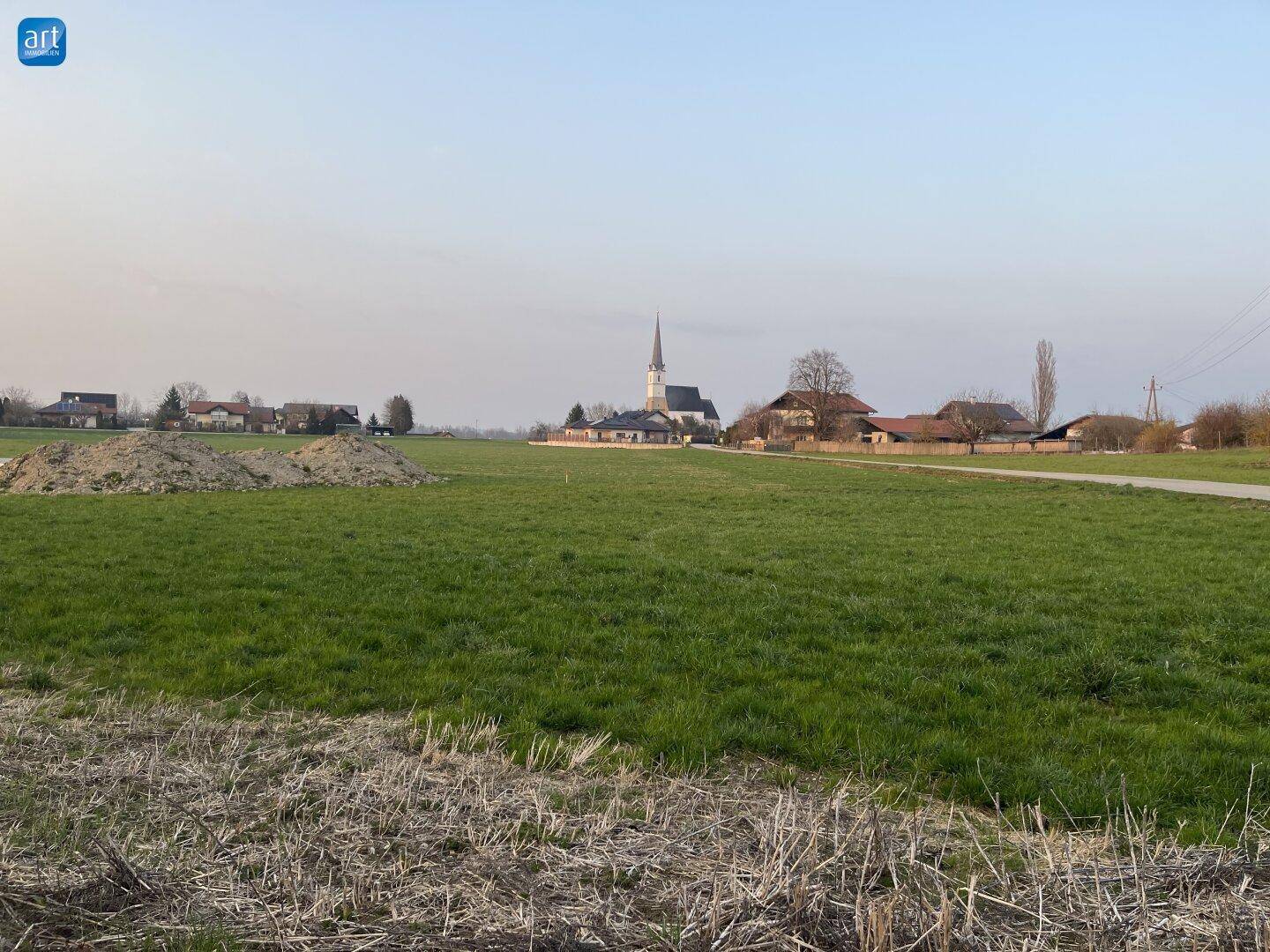 Weitläufige grüne Landschaft mit einem Dorf und Kirchturm am Horizont unter weitem Himmel.