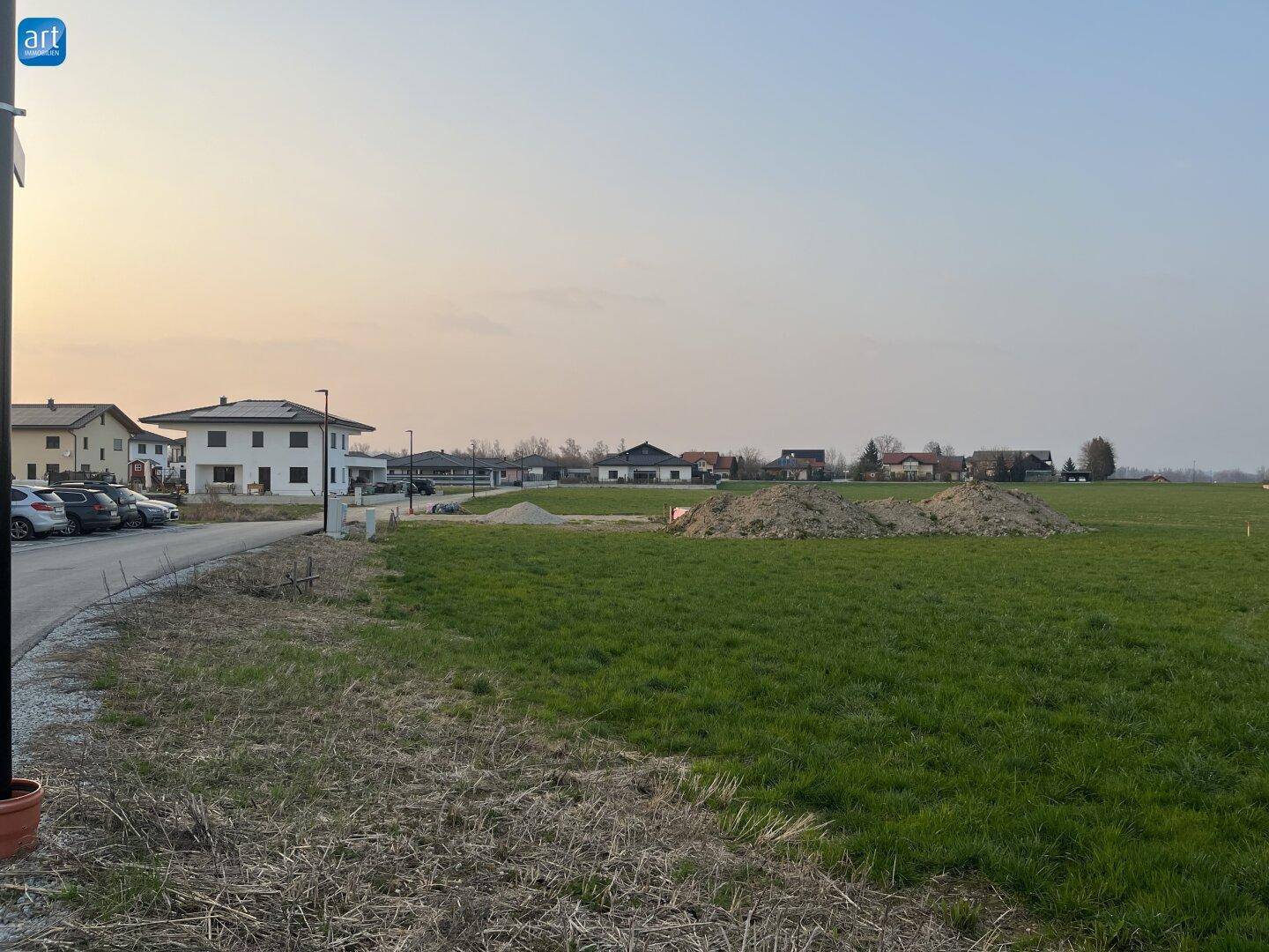 Grünfläche mit Zufahrtsweg und Blick auf neuere Wohnhäuser in ländlicher Umgebung bei Dämmerung.