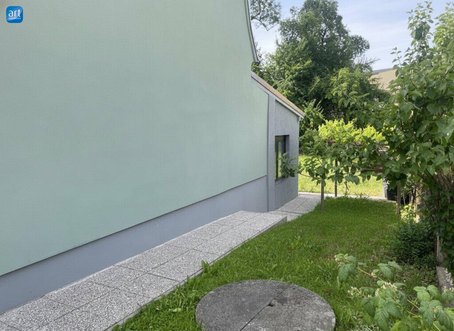 Seitenansicht des Hauses mit gepflastertem Weg, Rampe und grüner Bepflanzung im Gartenbereich.