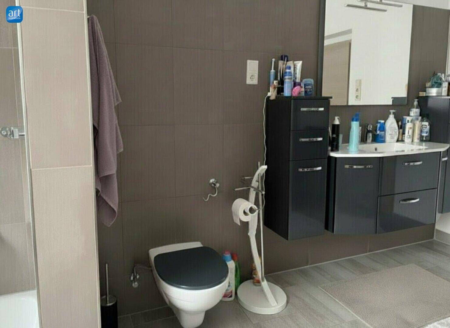 Modernes Badezimmer mit Waschtisch, Spiegel, Toilette und grauen Fliesen an Wänden und Boden.