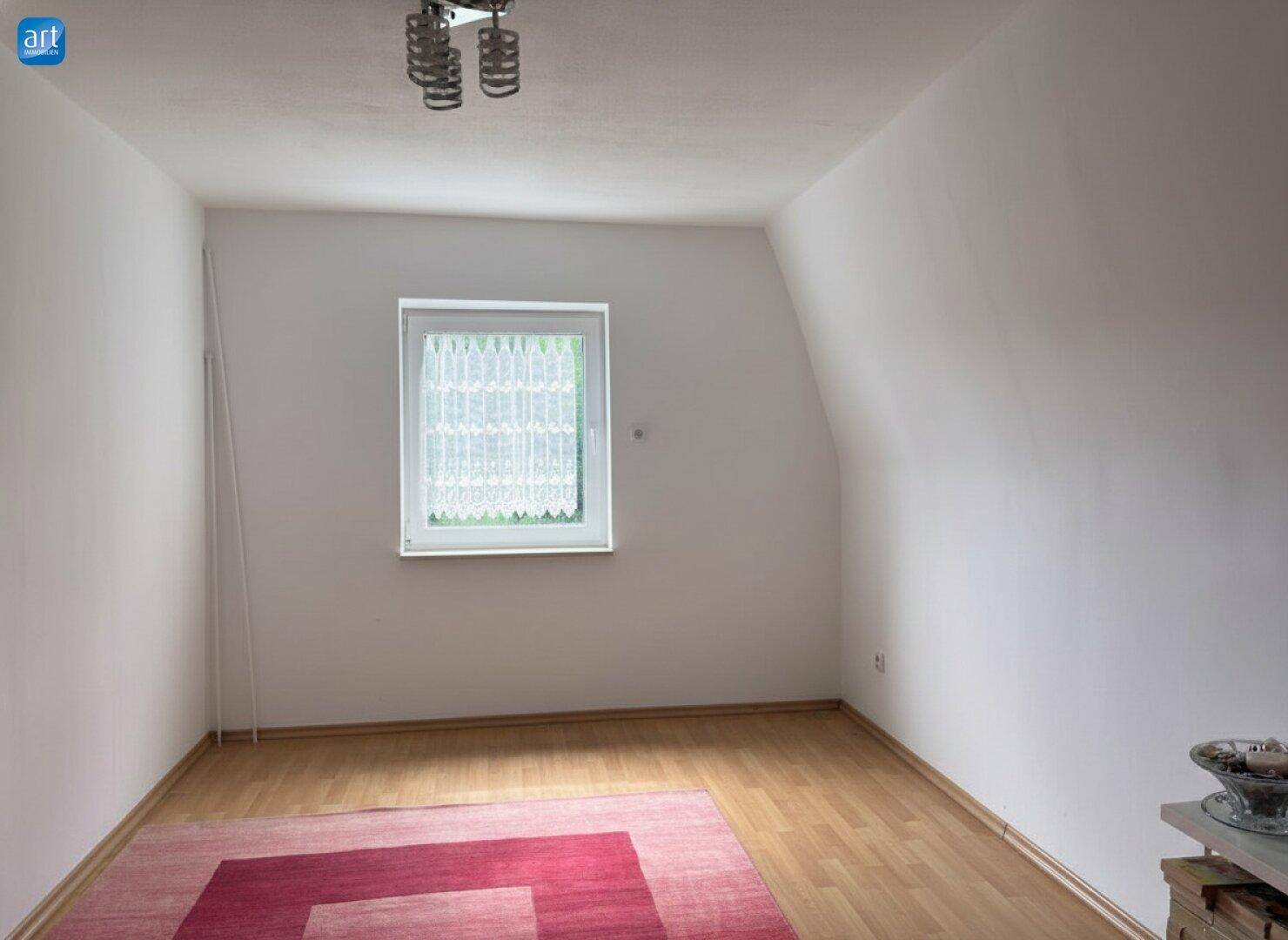 Helles Schlafzimmer mit Dachschräge, Holzboden und einem Fenster mit Vorhang, unmöbliert.