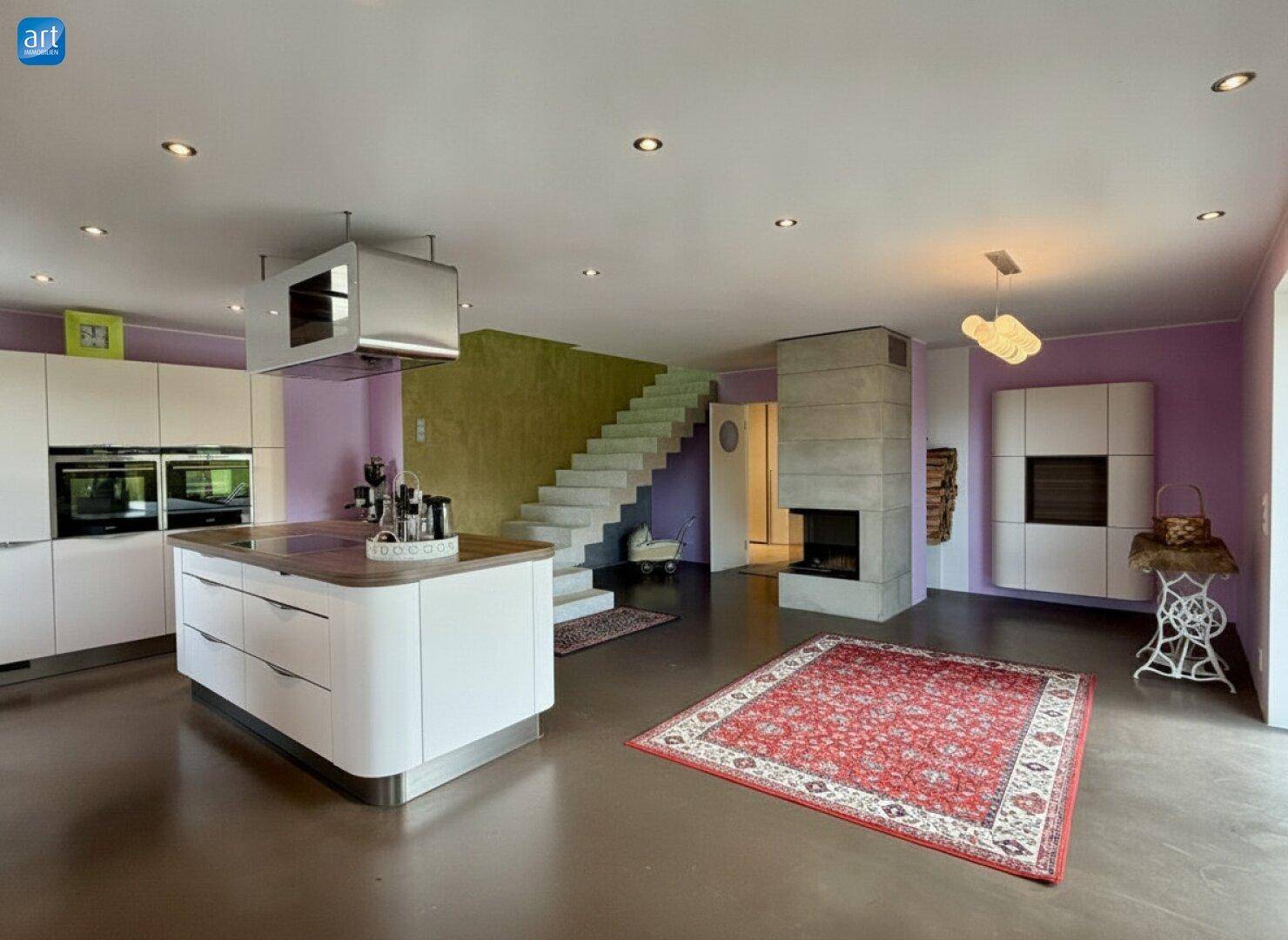 Offener Wohnbereich mit moderner Küche, Kamin und Treppe zum Obergeschoss, beleuchtet von Deckenspots.