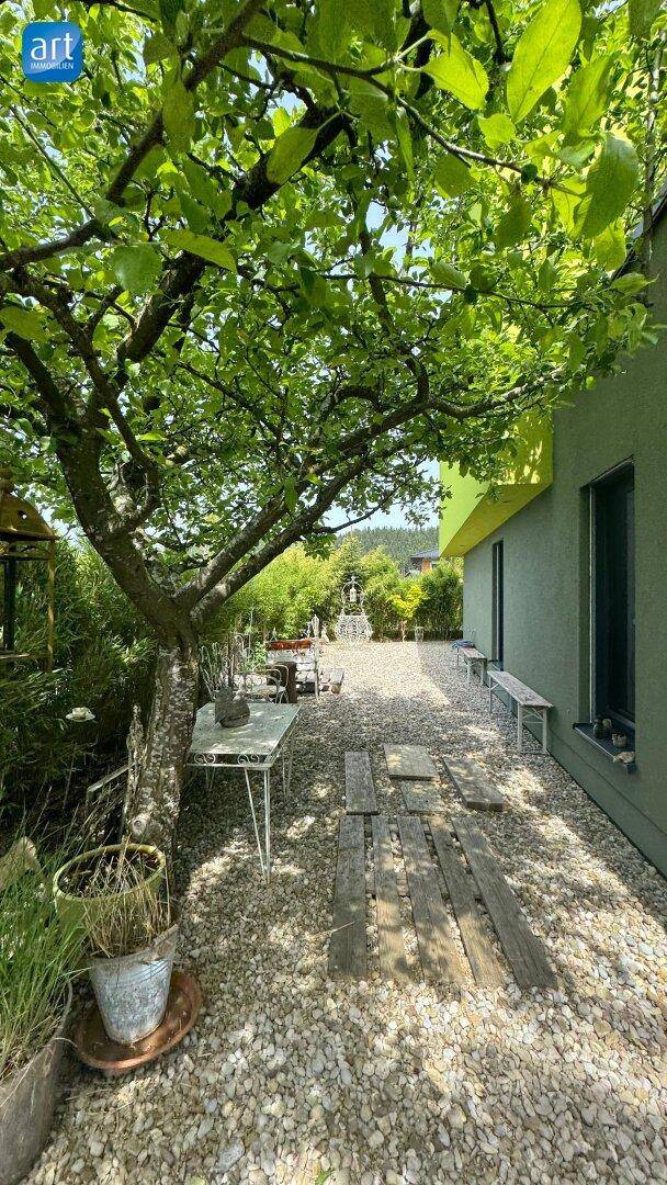 Kiesgarten mit schattenspendendem Baum, Sitzgelegenheiten und einem Pfad aus Holzbohlen.
