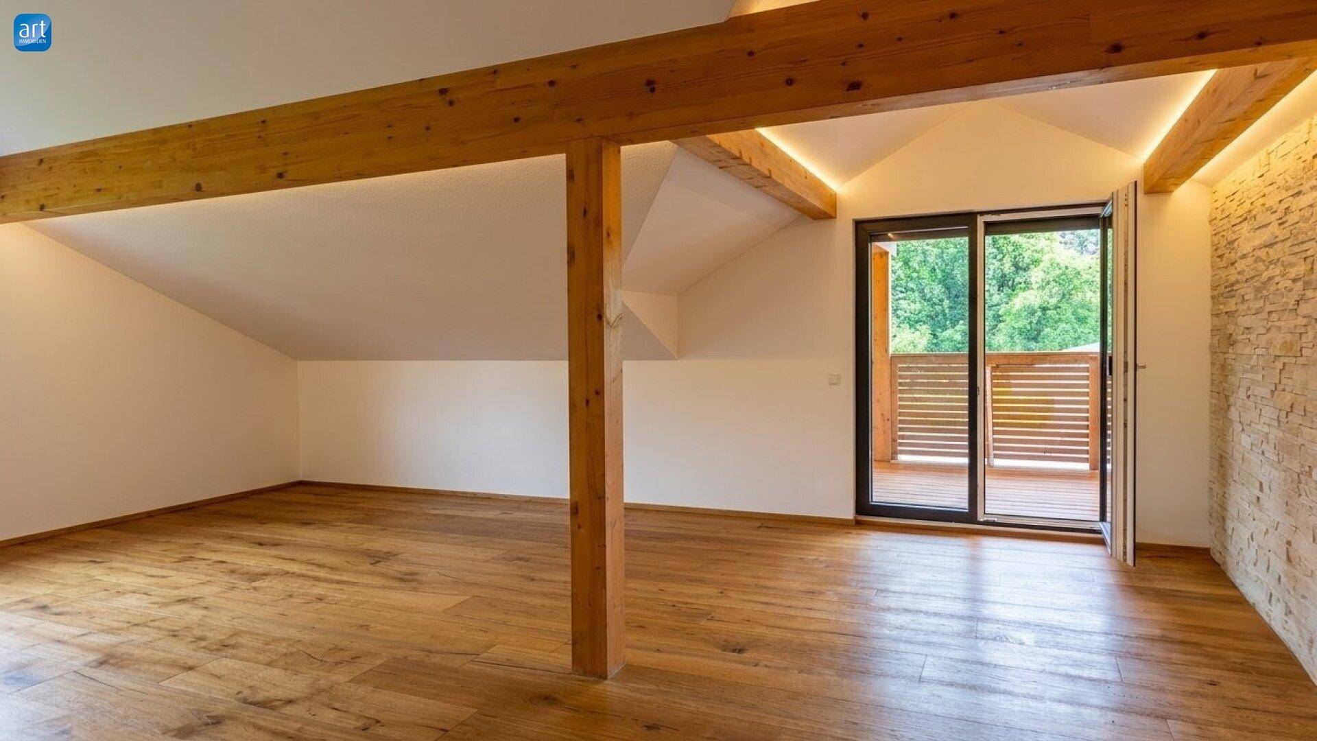 Geräumiger Dachgeschossraum mit sichtbaren Holzbalken, Holzboden und Zugang zu einem Balkon.