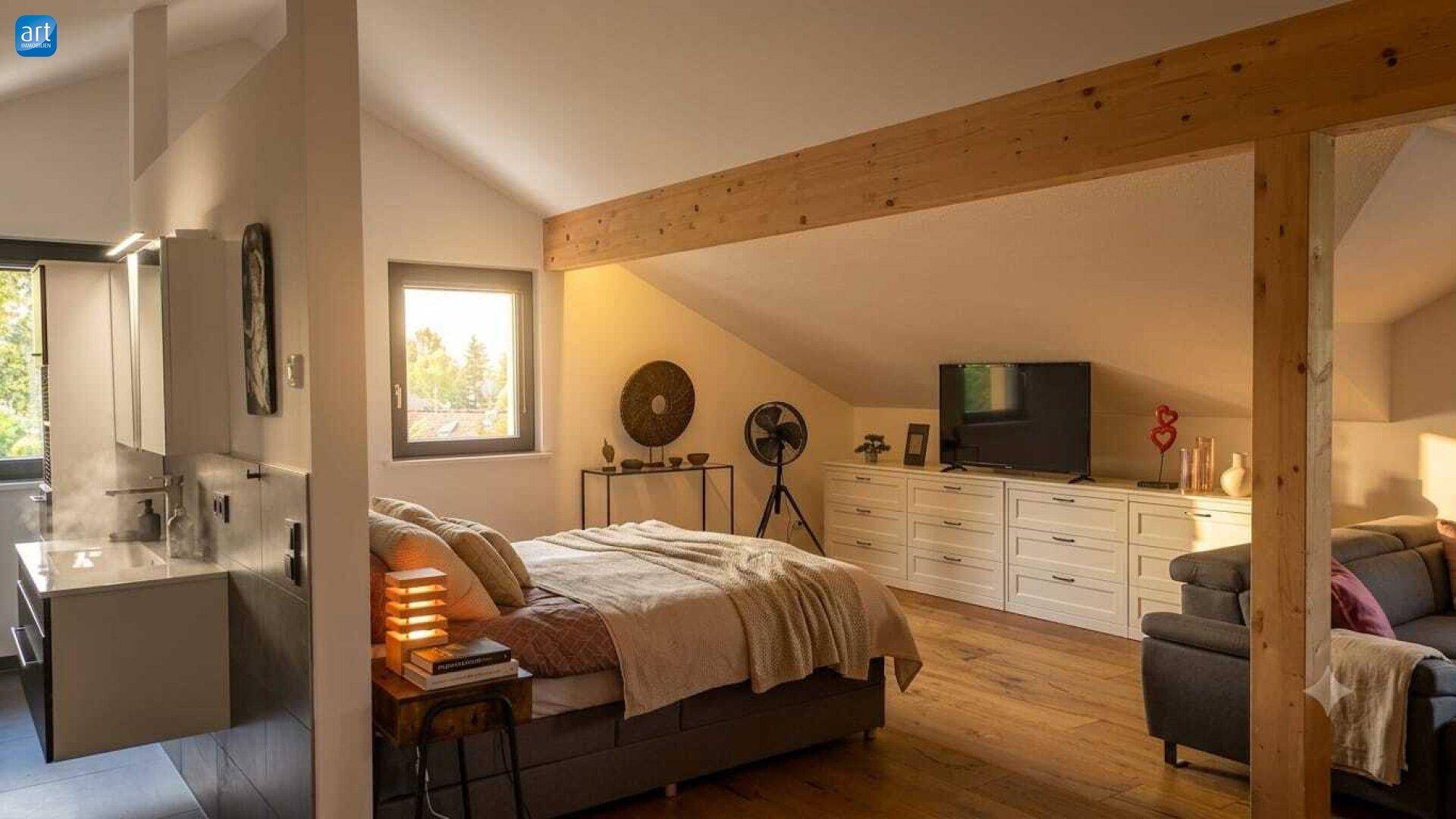 Offener Wohnbereich im Dachgeschoss mit Holzbalken, Bett und Fernseher, hell beleuchtet.