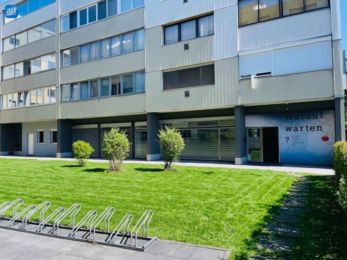 Moderne Gebäudefassade mit großen Fenstern, Fahrradständern und einer grünen Rasenfläche im Vordergrund.