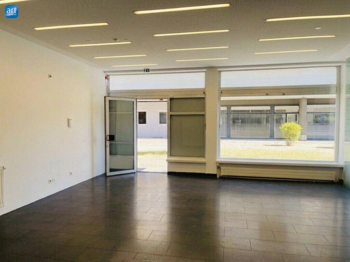 Heller, leerer Raum mit dunklem Fliesenboden, großen Fenstern und moderner Deckenbeleuchtung.
