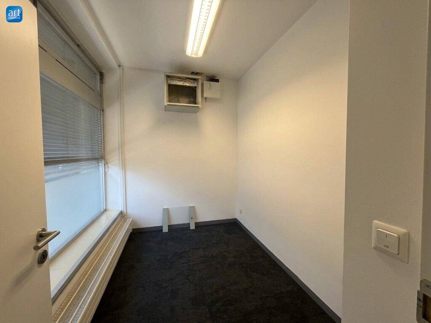Kleiner Raum mit dunklem Teppichboden, Fenster mit Jalousien und sichtbarer Klimaanlage.