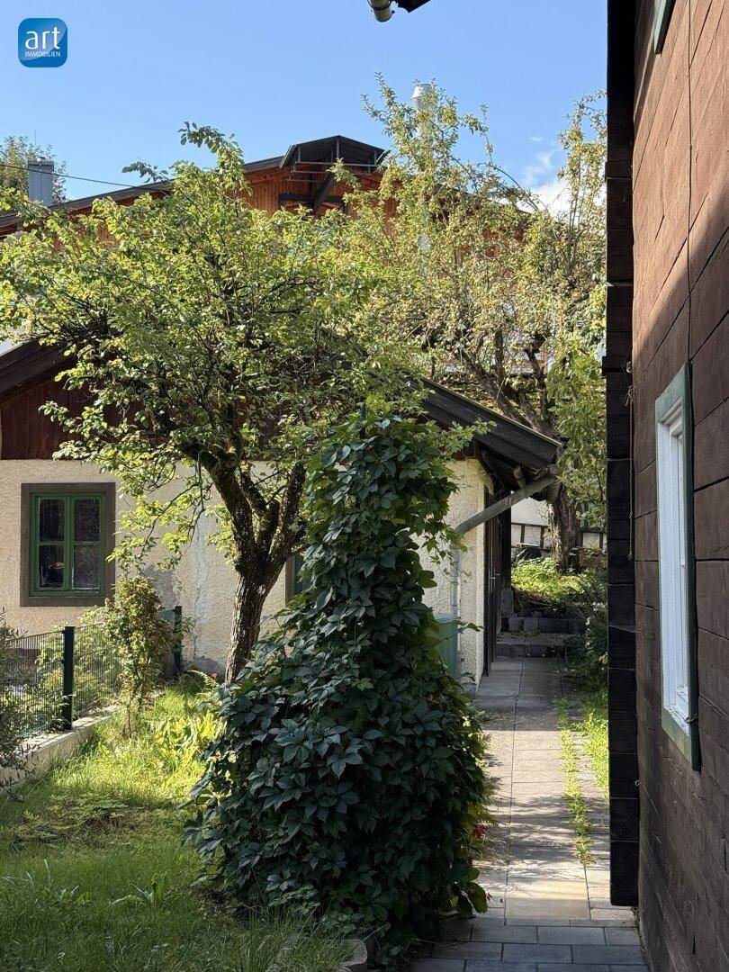 Seitlicher gepflasterter Weg neben dem Haus mit üppiger Vegetation und Nebengebäude.