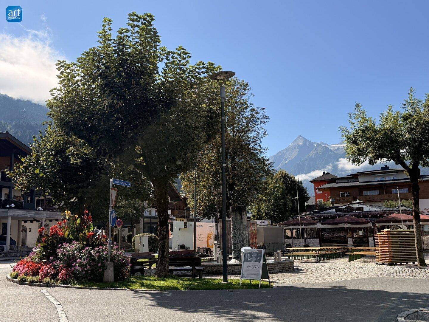Malerischer Stadtplatz mit Bäumen, Blumenbeeten und Bergblick im Hintergrund.