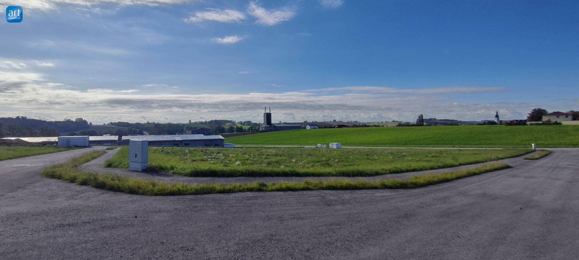 Offene Landschaft mit weiten grünen Flächen, einer Straße und Gebäuden in der Ferne unter blauem Himmel.