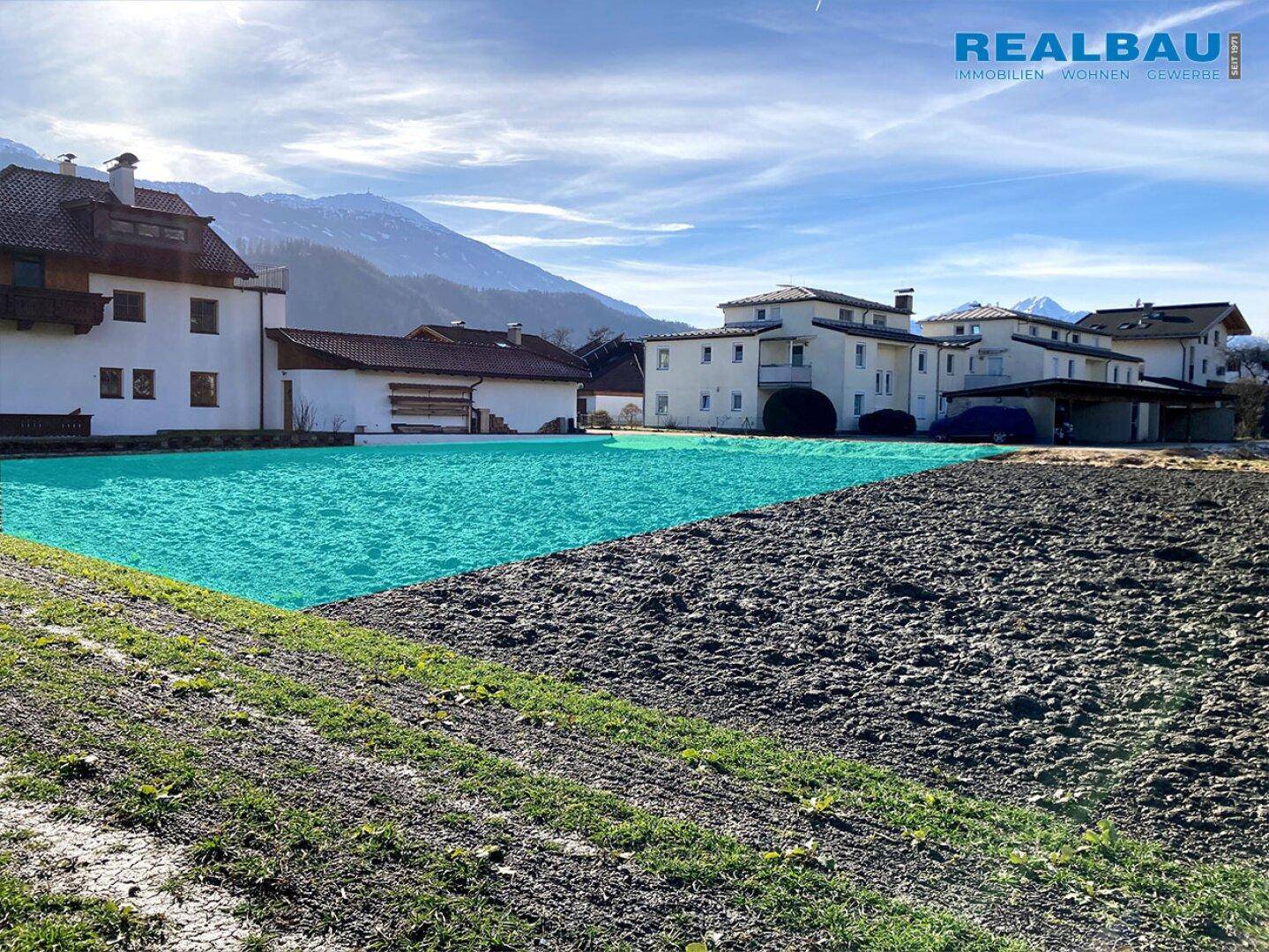 Blick auf das unbebaute Grundstück mit umliegenden Häusern und Alpenpanorama im Hintergrund unter blauem Himmel.