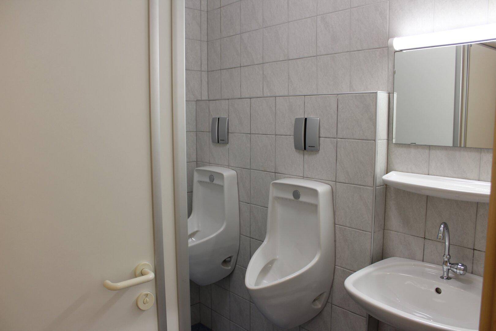 Herrenwaschraum mit zwei Urinalen, einem Waschbecken und einem Spiegel, alles sauber gefliest.
