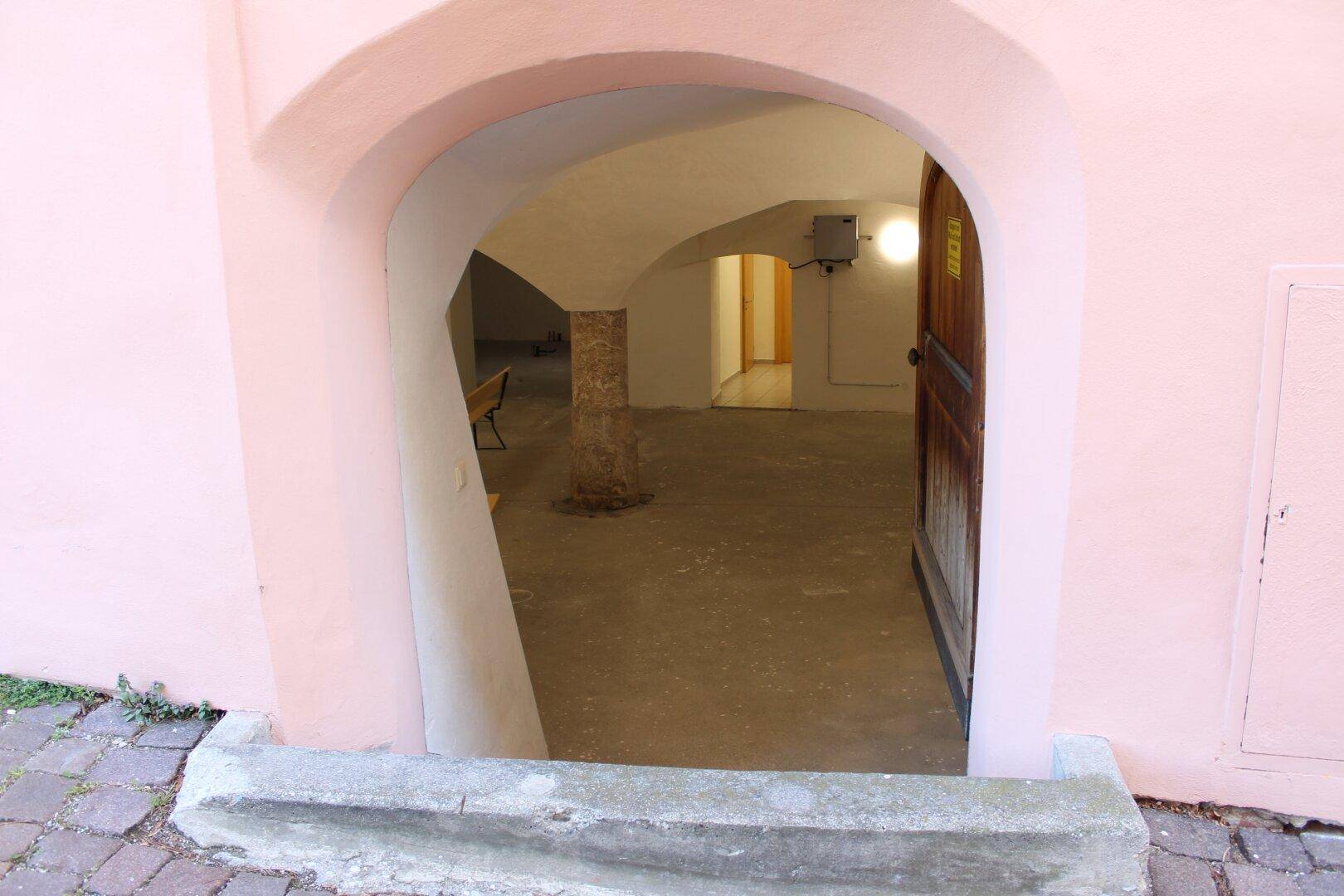 Blick durch einen gewölbten Eingang in einen geräumigen Kellerbereich mit Tonnengewölbe und einem Steinpfeiler.
