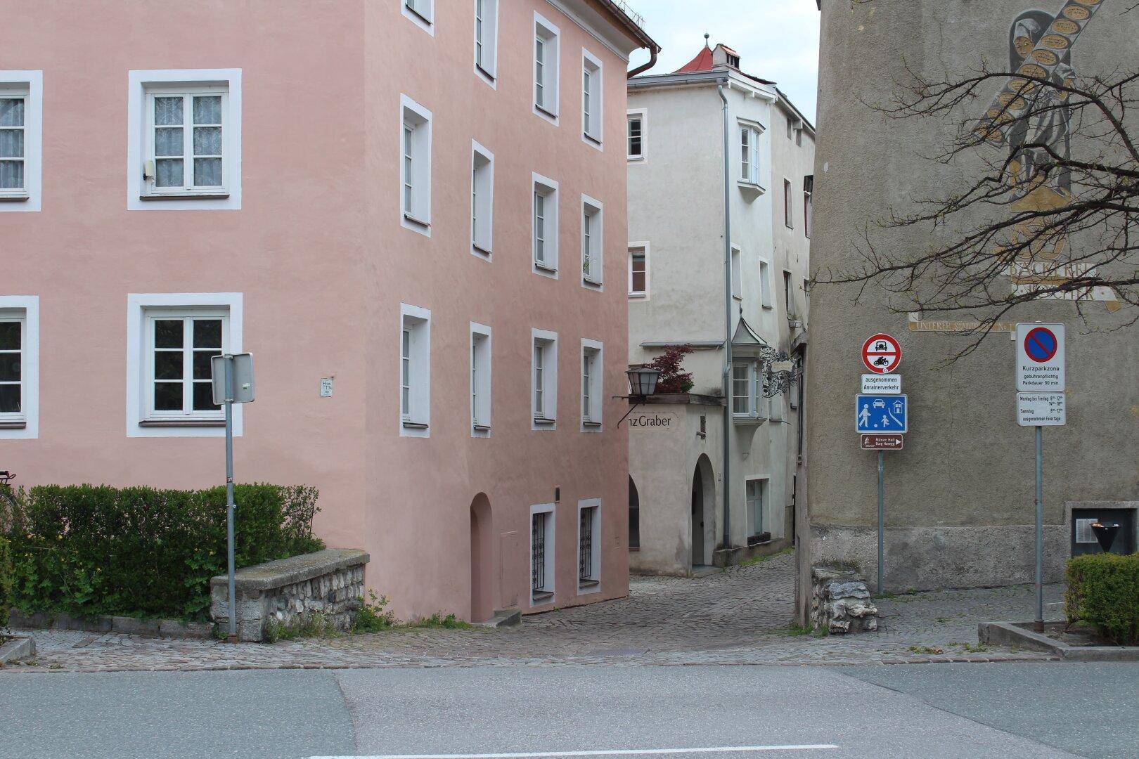 Straßenansicht mit historischen Gebäuden, darunter ein rosafarbenes Haus, und verschiedenen Verkehrsschildern.