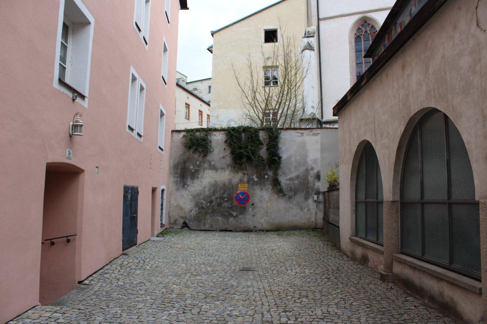 Historischer Innenhof mit Kopfsteinpflaster, umgeben von alten Gebäuden und einer hohen, bewachsenen Mauer.