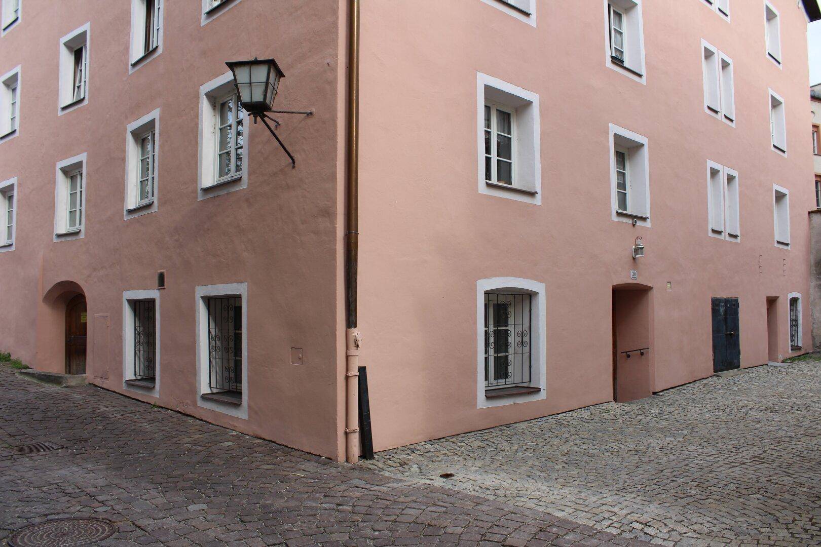 Eckansicht eines historischen Gebäudes mit rosafarbener Fassade, weißen Fenstern und Kopfsteinpflaster im Vordergrund.