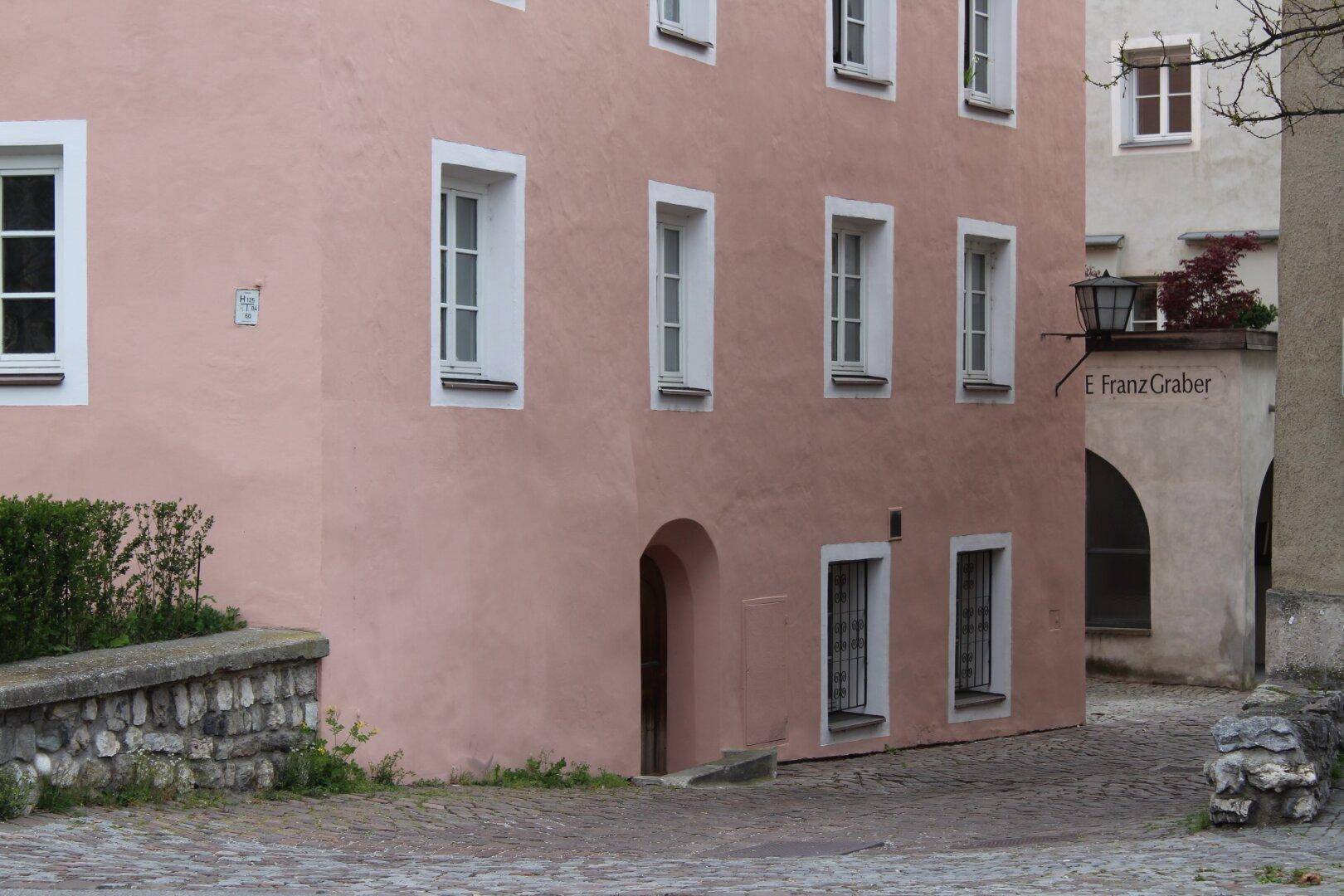 Rosafarbenes Gebäude mit weißen Fenstern und einem kleinen Busch, an einer Kopfsteinpflasterstraße gelegen, historischer Charme.