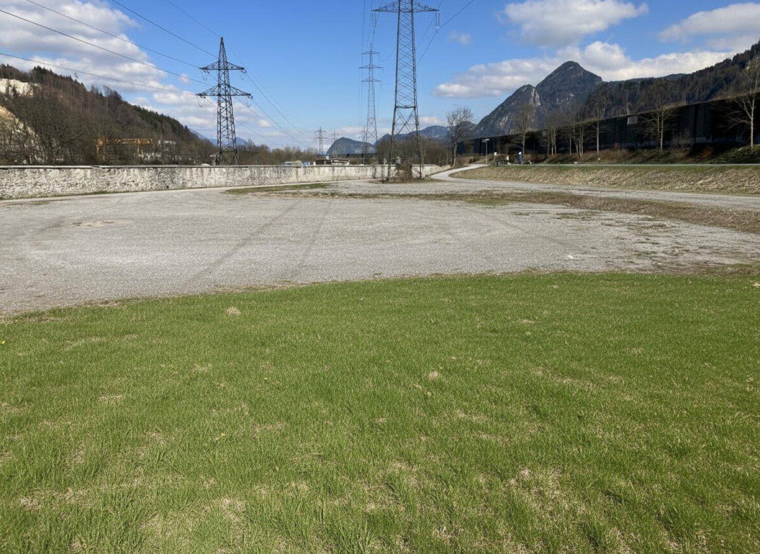 Große Schotterfläche mit Grasstreifen, umgeben von Bergen und Stromleitungen unter blauem Himmel.