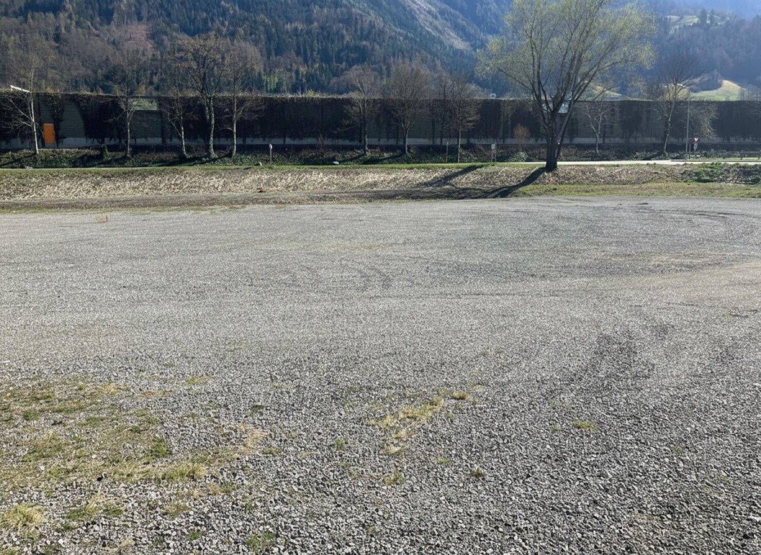 Große Schotterfläche mit Bäumen und einer Lärmschutzwand im Hintergrund, umgeben von Natur.