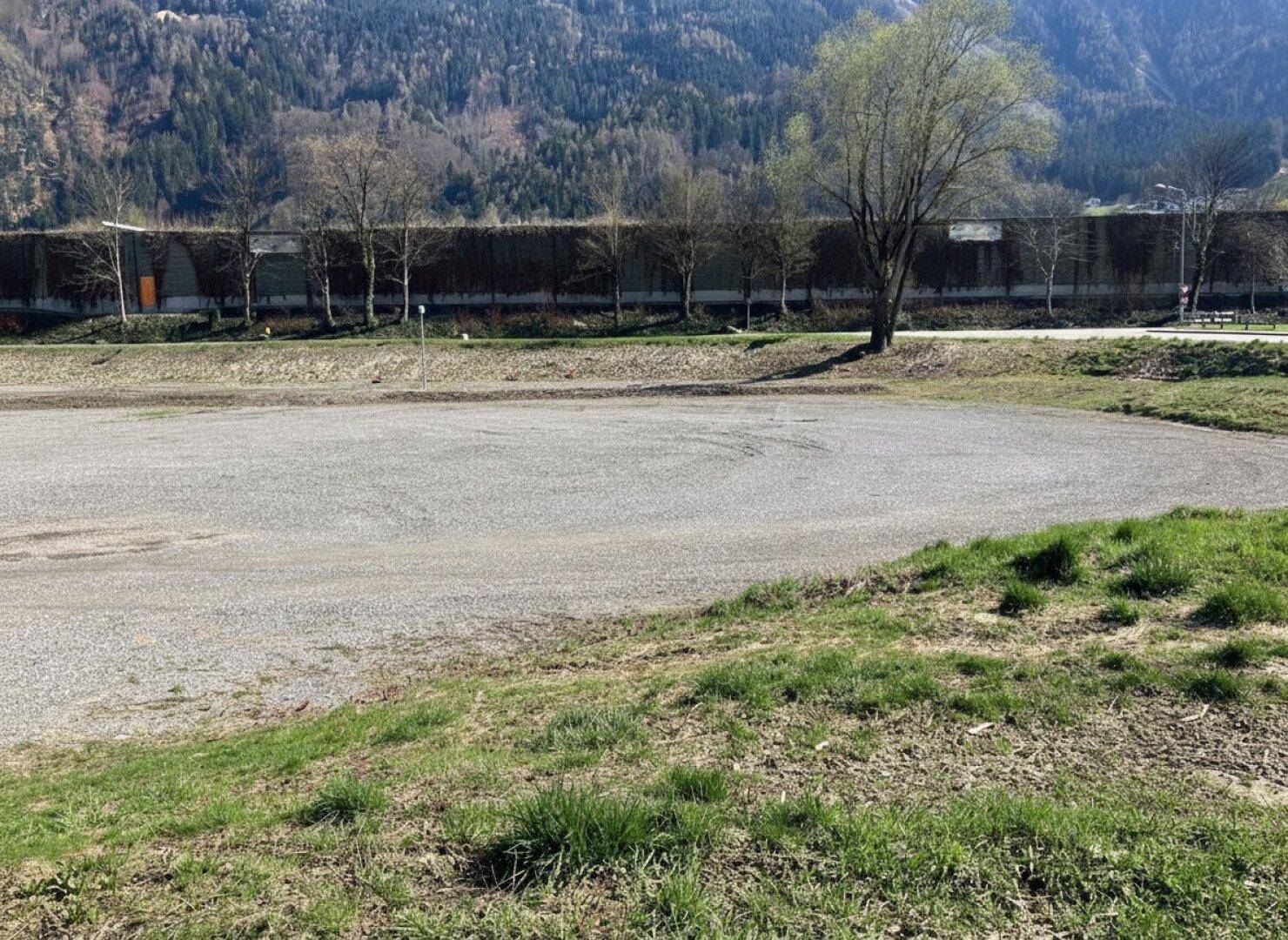 Weitläufiges Schottergrundstück mit Grasflächen und Bäumen, umgeben von einer bergigen Landschaft.