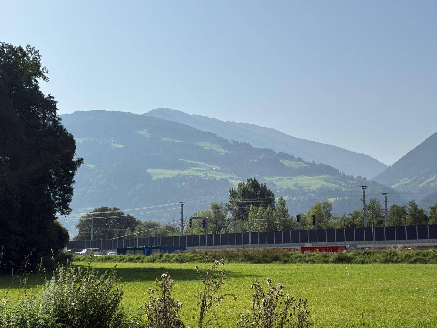 Weitläufiger Blick über grüne Felder und eine Lärmschutzwand hin zu einer ausgedehnten Bergkette unter klarem Himmel.