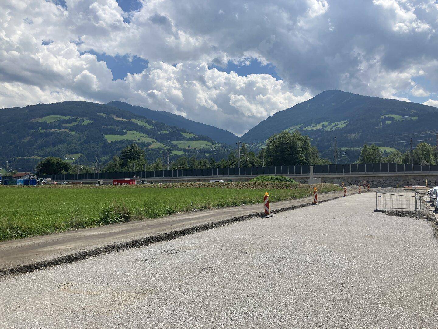Baustellenbereich mit neuem Schotterweg, umgeben von grünen Feldern und Bergen im Hintergrund unter bewölktem Himmel.