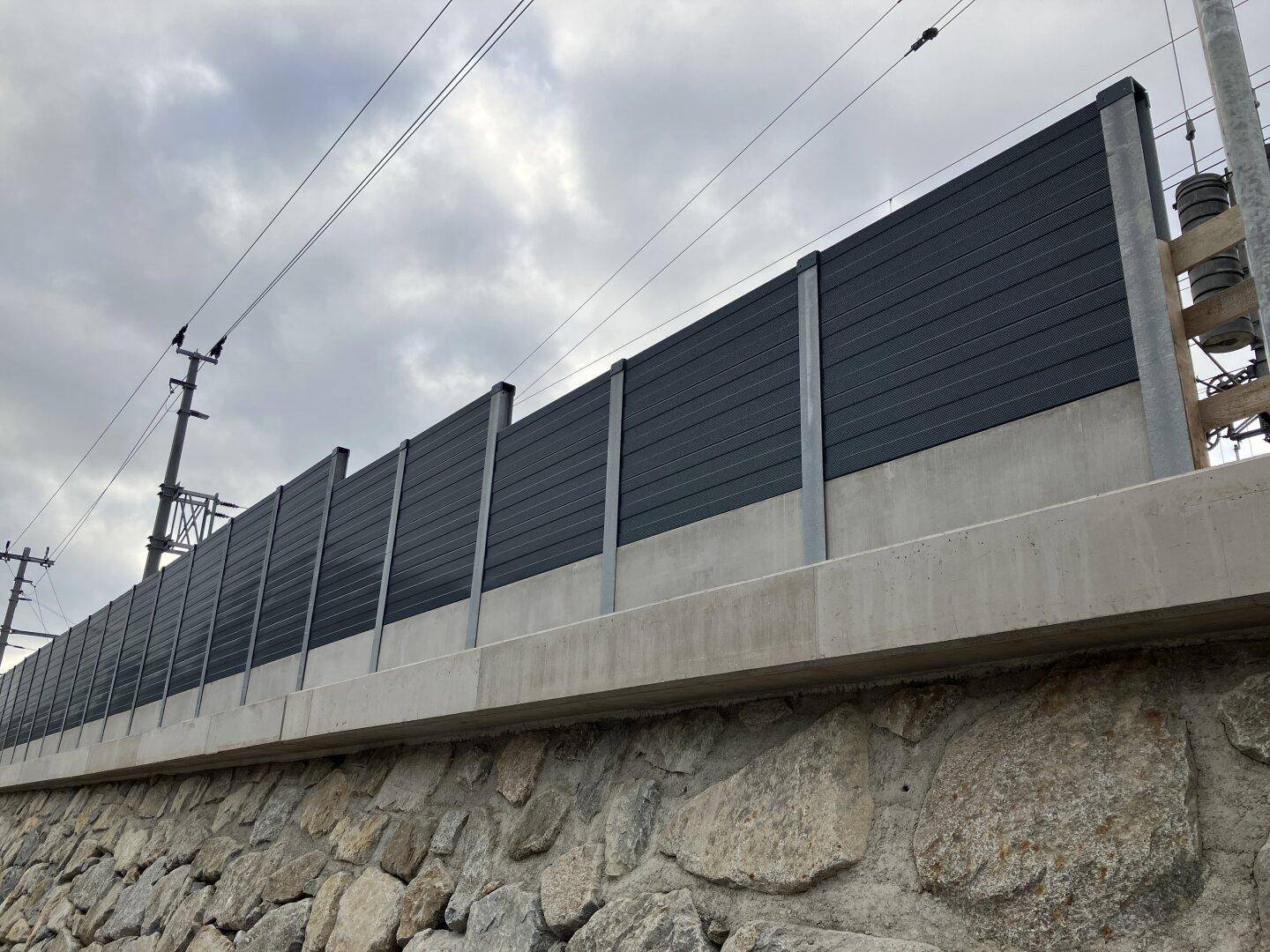 Detailansicht einer modernen Lärmschutzwand aus Beton und dunklen Paneelen auf einem Natursteinsockel.
