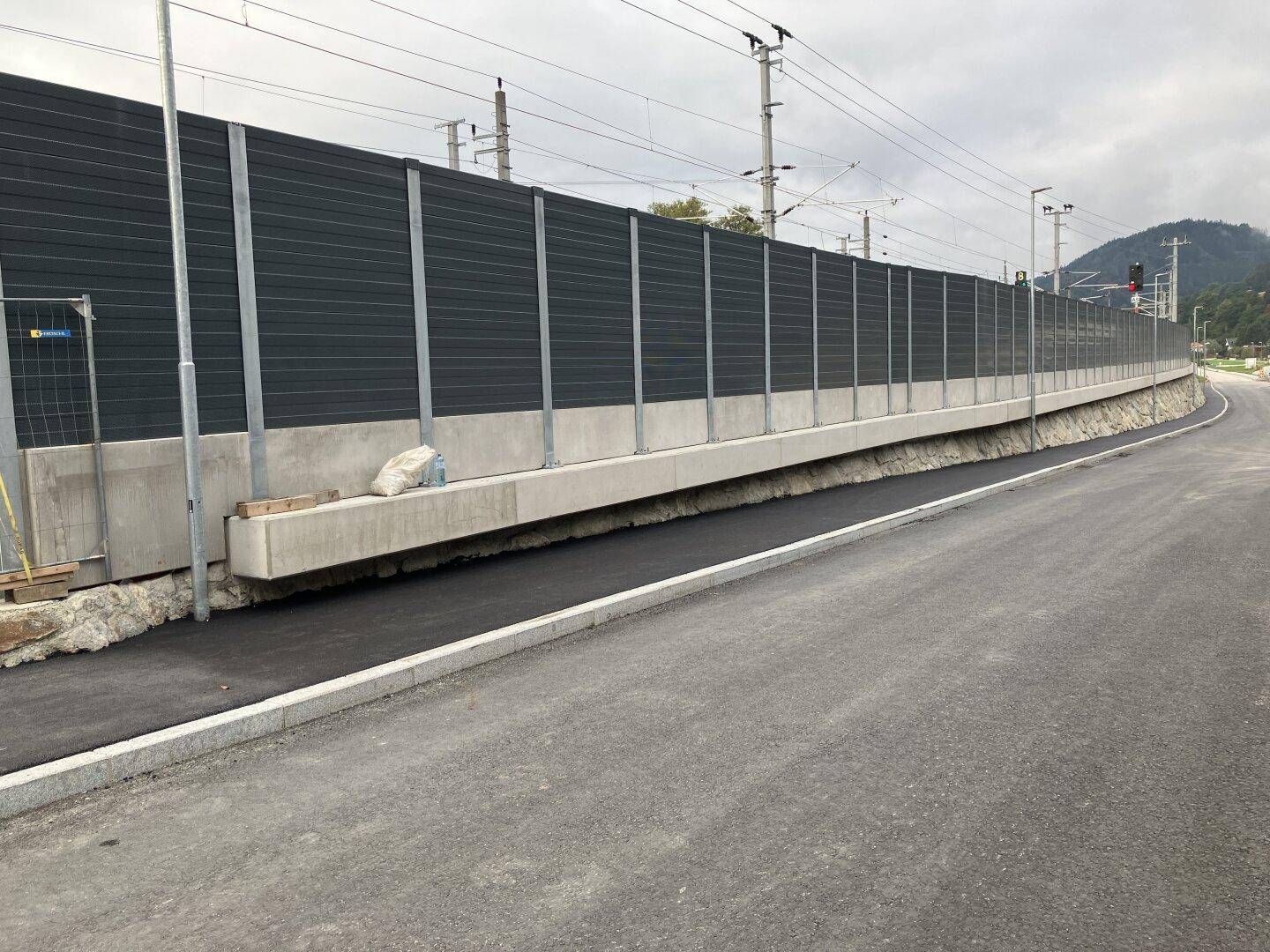 Lange Lärmschutzwand entlang einer neu asphaltierten Straße mit Bürgersteig und angrenzendem Grünstreifen.