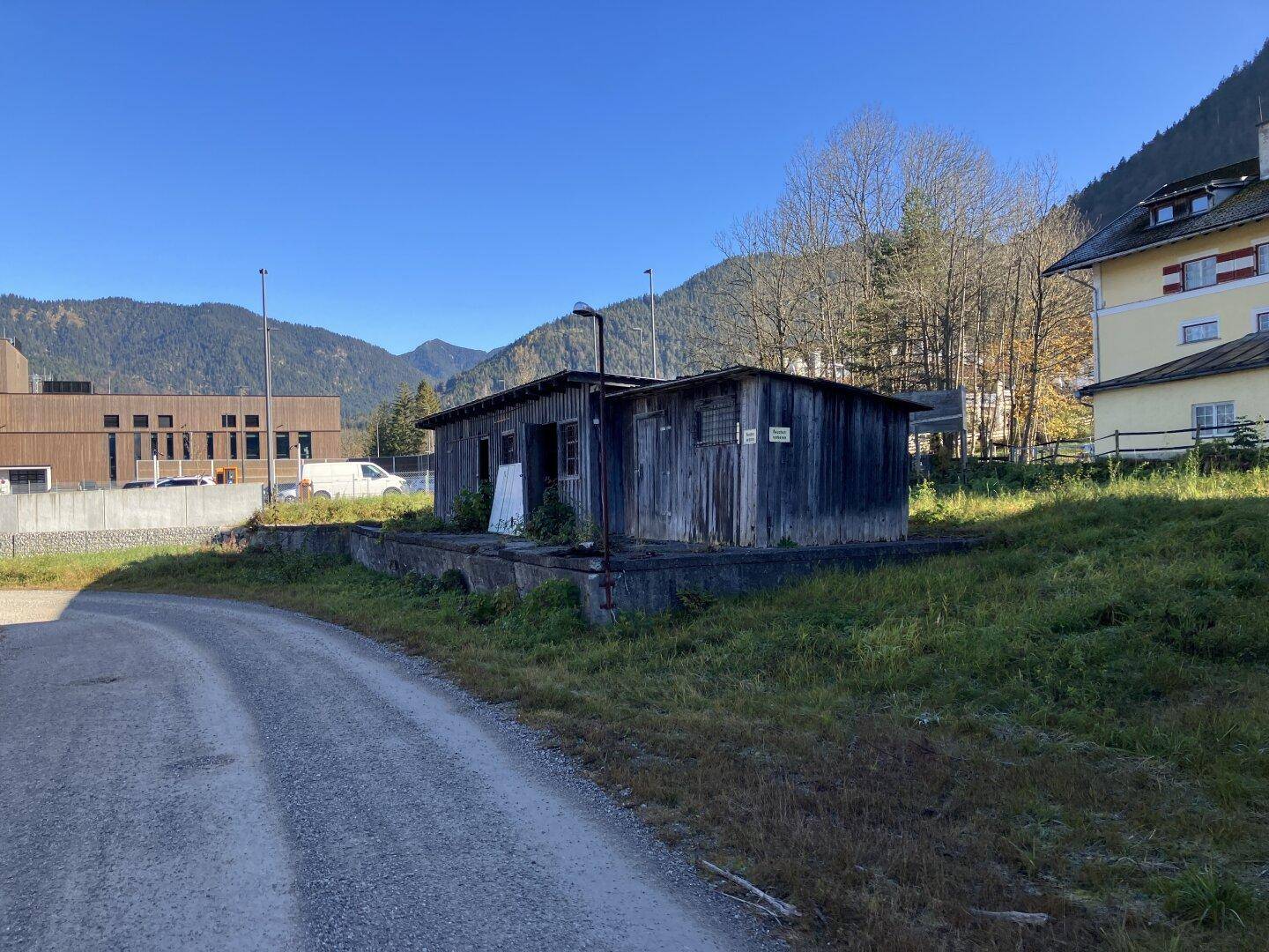 Kleines, verwittertes Holzgebäude neben einem Schotterweg mit Bergen und einem modernen Gebäude im Hintergrund.