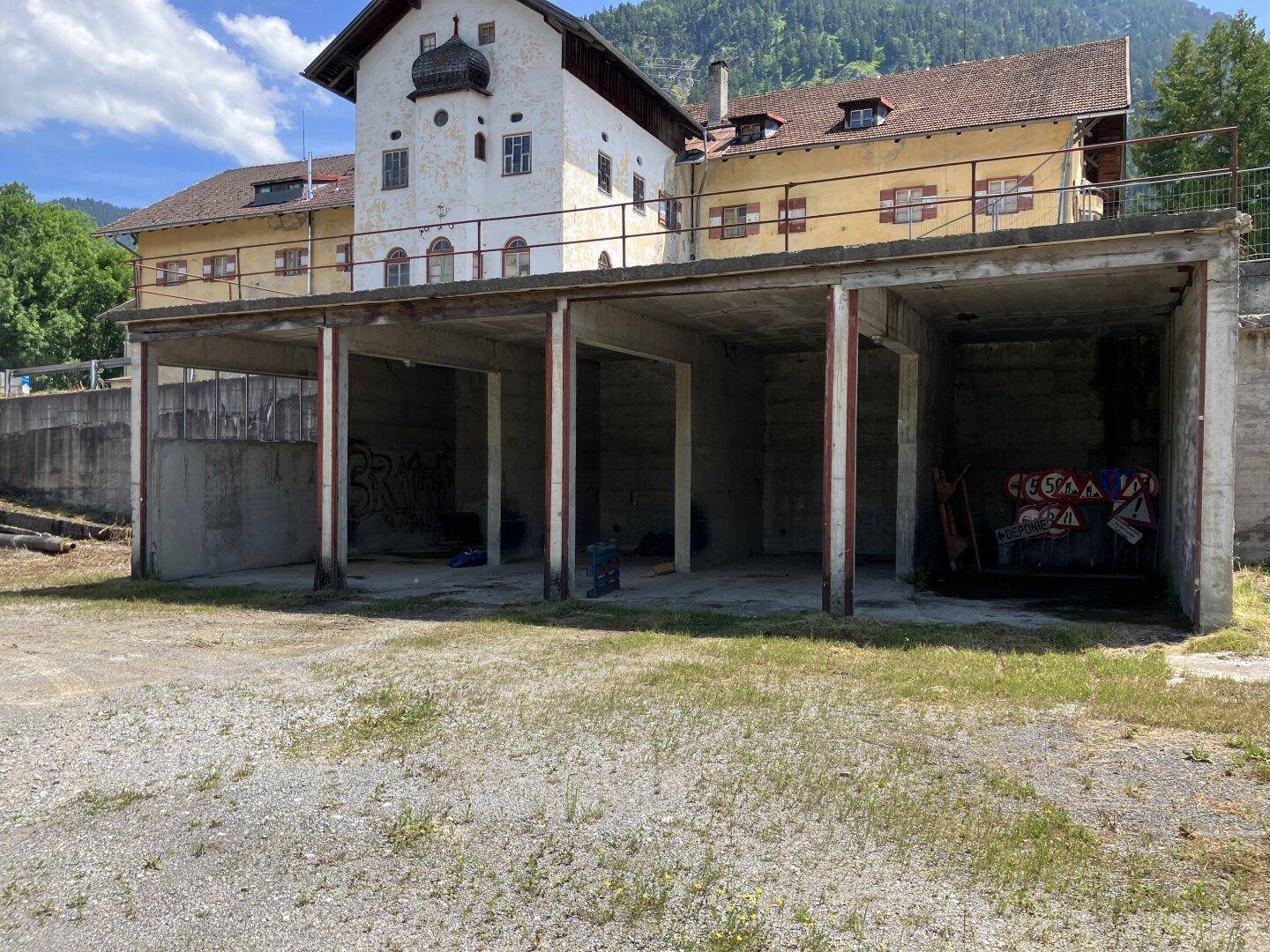 Offene Betonkonstruktion mit mehreren Pfeilern vor einem historischen Gebäude und grünen Bergen.