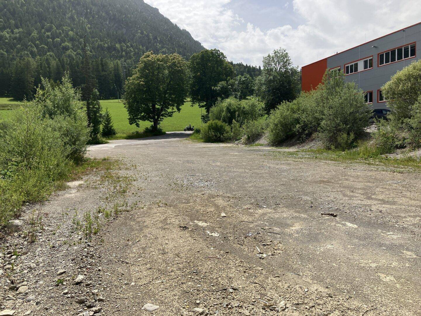 Schotterfläche mit umgebender Vegetation und einem Blick auf grüne Hügel und Berge im Hintergrund.