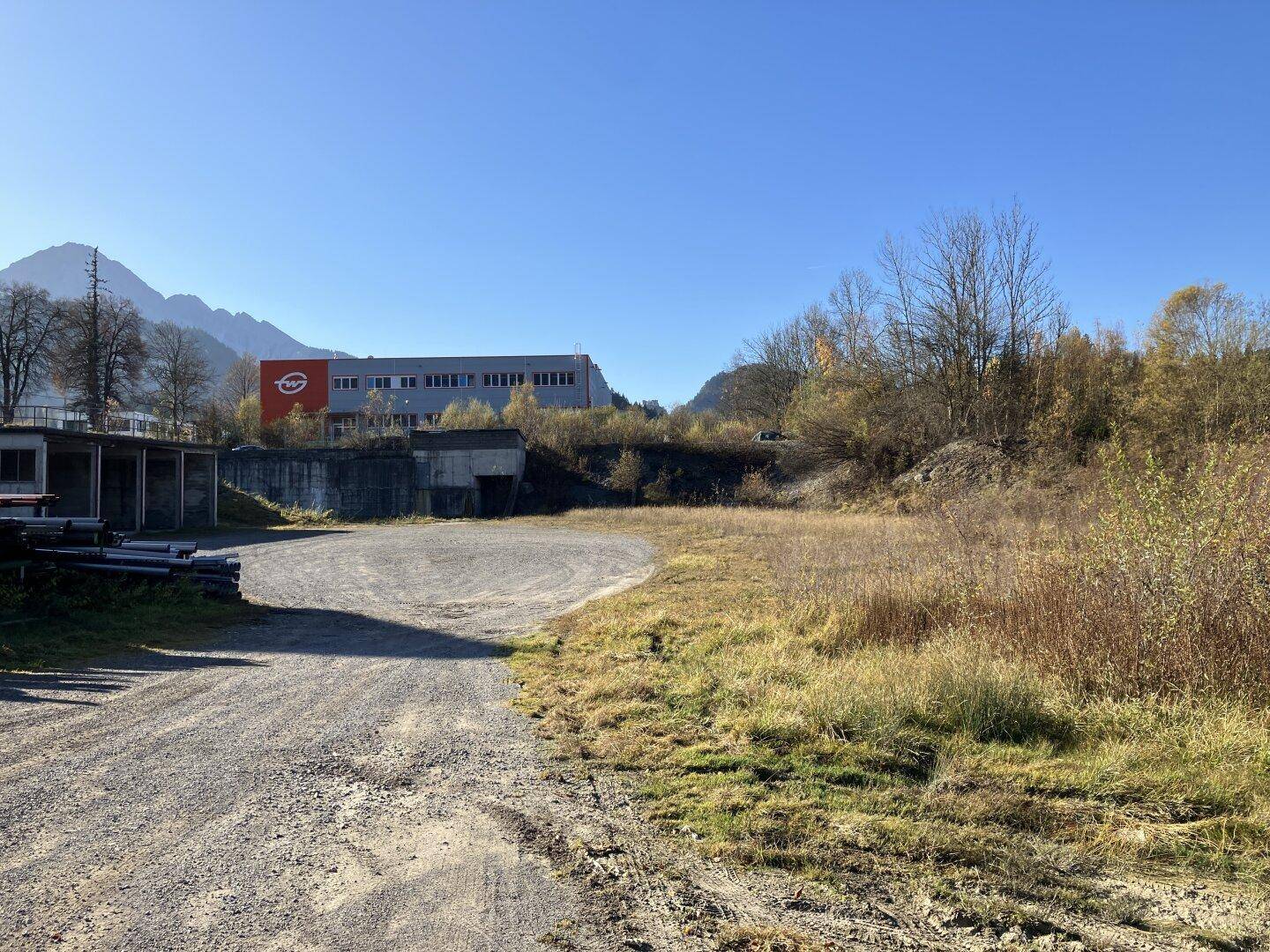 Großes, offenes Grundstück mit trockenem Gras, einem Industriegebäude und Bergen im Hintergrund an einem sonnigen Tag.