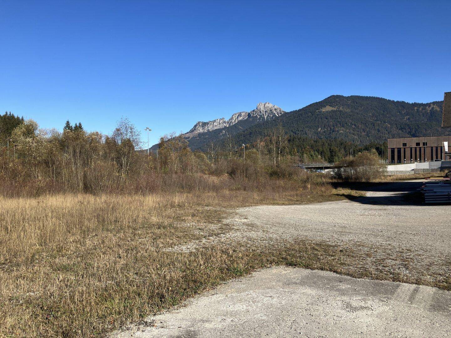 Offenes Grundstück mit trockener Vegetation und Schotterflächen, umgeben von bewaldeten Bergen unter klarem Himmel.