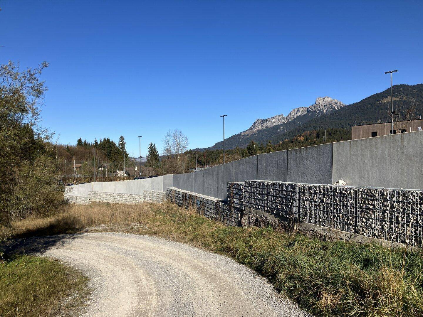 Lange Betonmauer und Gabionenwand entlang eines Schotterwegs, der zu einem Gebäude führt, mit Bergen im Hintergrund.