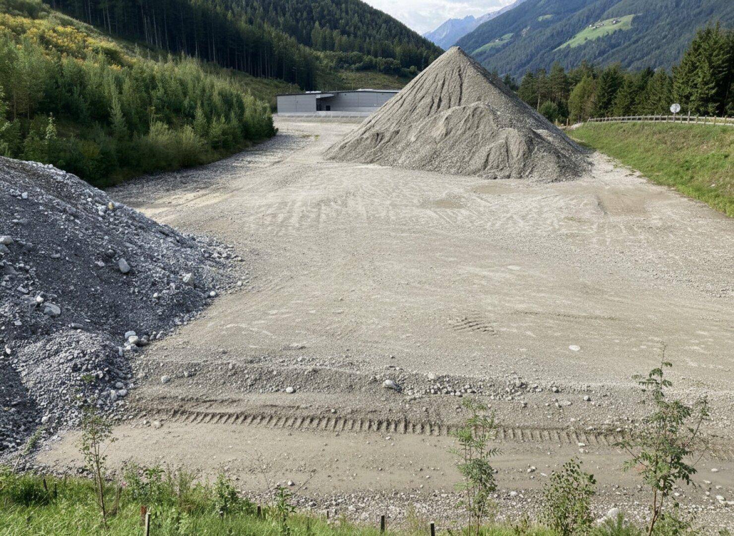 Weitläufiges Schottergelände mit großen Kieshaufen und einem Gebäude im Hintergrund, umgeben von bewaldeten Bergen.