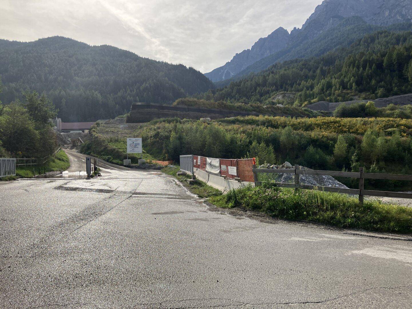 Nasse Asphaltstraße führt zu einem Industriegebiet in einem Tal, umgeben von bewaldeten Bergen unter einem bewölkten Himmel.