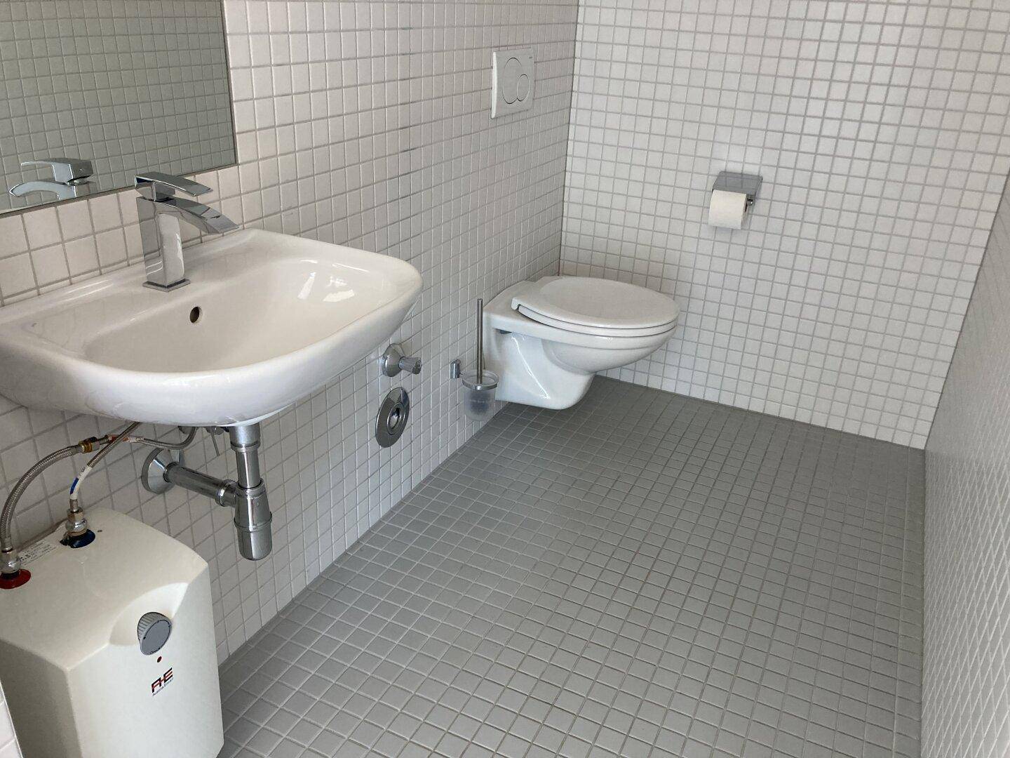 Helles Badezimmer mit kleinen weißen Fliesen an den Wänden und grauen Bodenfliesen, ausgestattet mit Waschbecken und Toilette.