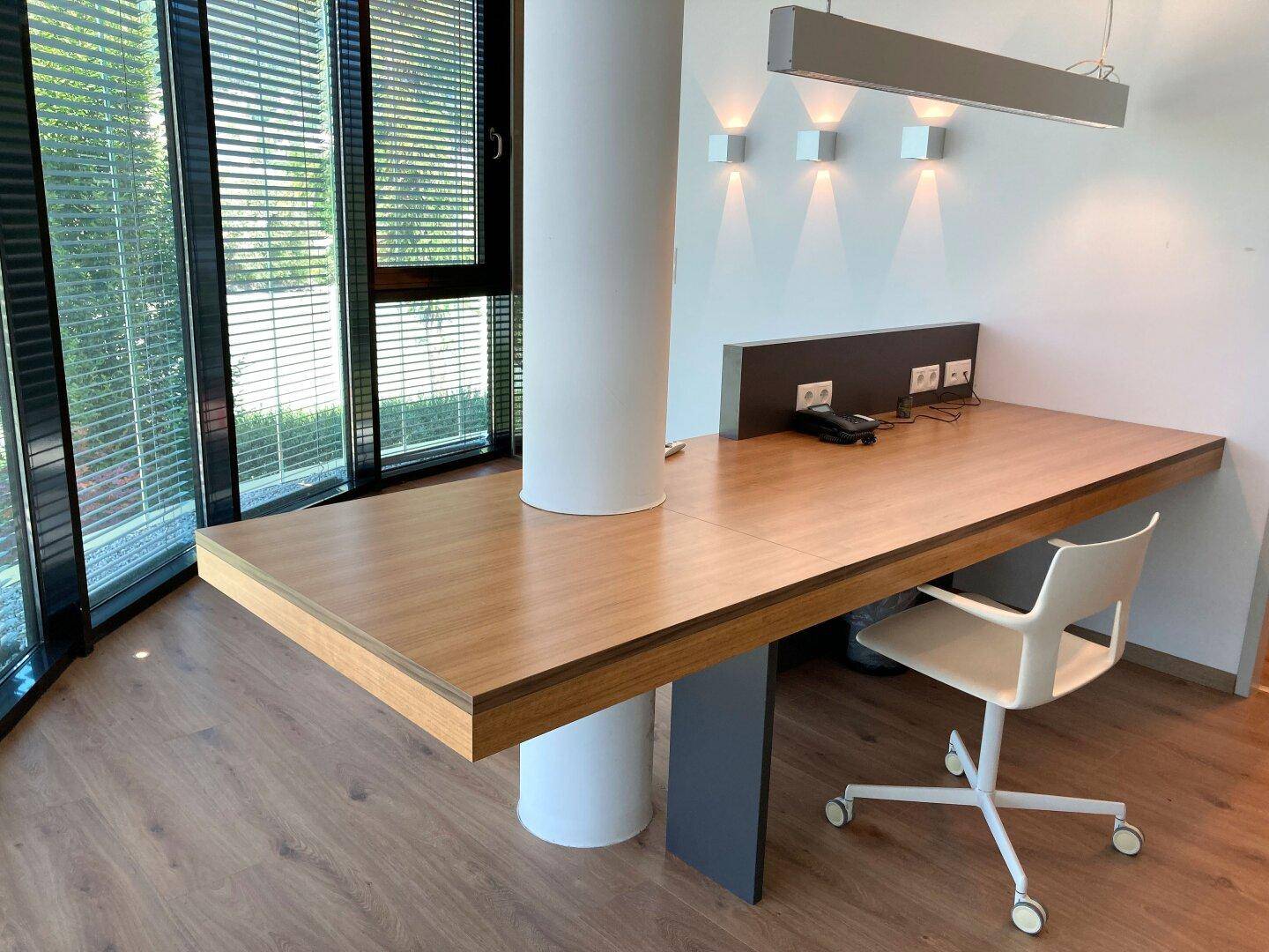 Moderner Arbeitsplatz mit integriertem Schreibtisch um eine Säule, großen Fenstern und Holzboden.