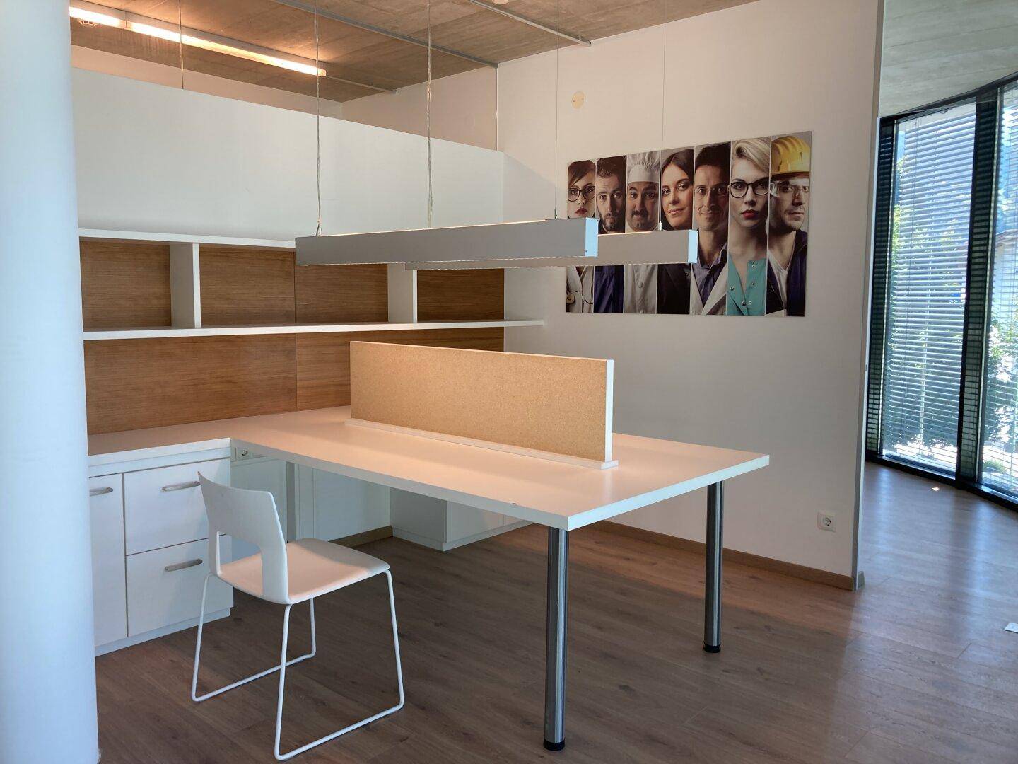 Offener Bürobereich mit mehreren Arbeitsplätzen, Regalen und großen Fenstern, die den Raum hell erscheinen lassen.